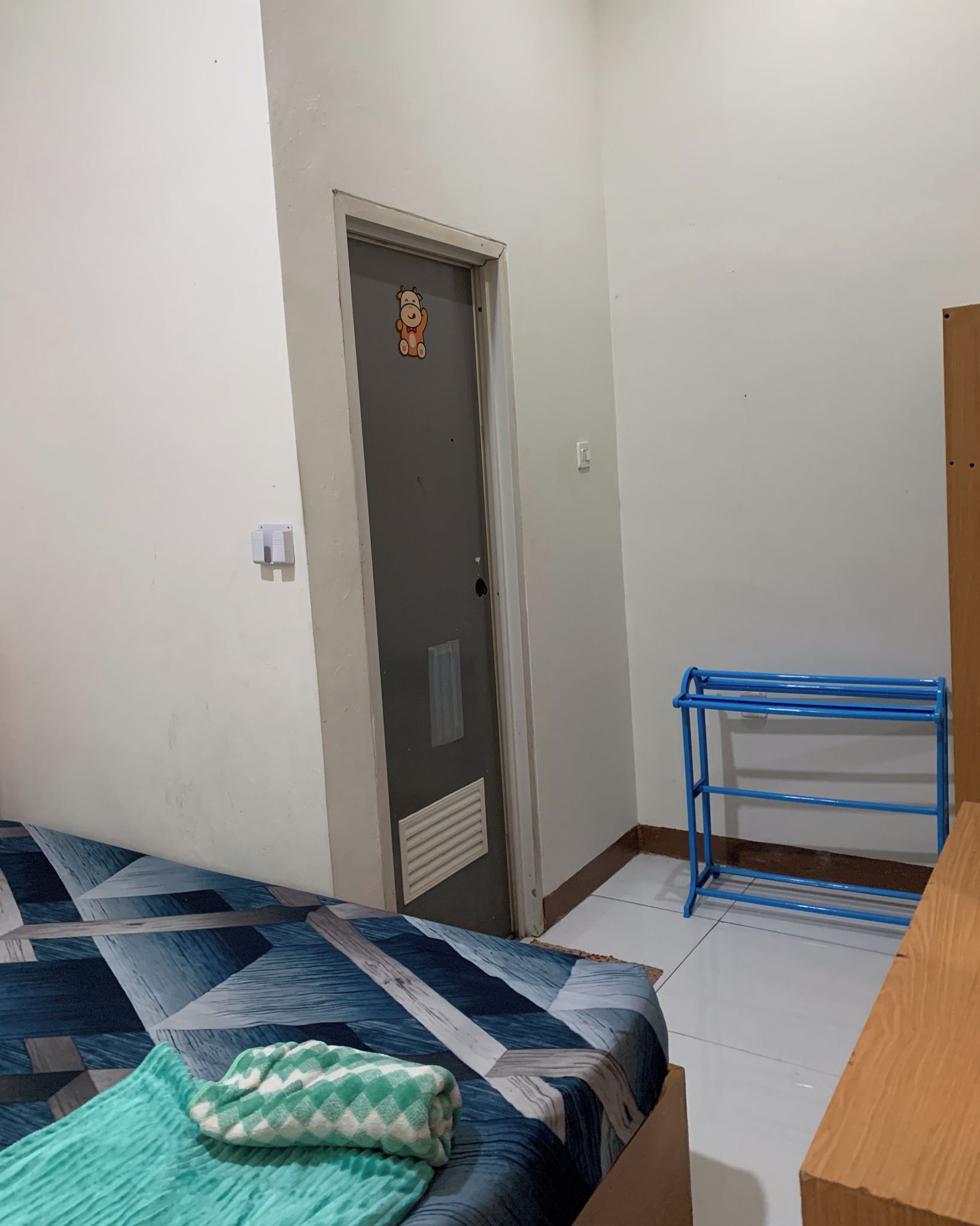 Una habitación pequeña y poco amoblada con una cama, un estante metálico azul y una puerta con un personaje de dibujos animados.