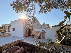 A white building with a stone facade, a tiled patio, and a small garden area with olive trees. - Trullo Ostro - Fable Retreats - Chic Puglia Style (Ostuni)