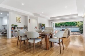 A modern dining area with a wooden table and four chairs, a bookshelf, and a view of the outdoors. - Casa Fern (Lennox Head)
