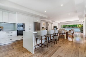 A modern kitchen with white cabinetry, a central island with bar stools, and a dining area with a view of the outdoors. - Casa Fern (Lennox Head)