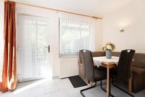 A room with a dining table, two chairs, a vase with flowers, a radiator, and a door with curtains. - Vrbo Property (Kellenhusen)