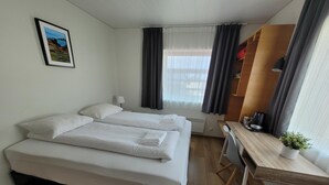 Ein Hotelzimmer mit zwei Betten, einem Schreibtisch, einem Stuhl, einem kleinen Tisch mit Pflanze, einem Bild an der Wand und einem Fenster mit Vorhängen.
