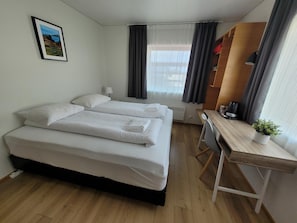 Et hotelværelse med to senge, et skrivebord, en stol, et lille bord med en plante, et billede på væggen og et vindue med gardiner.