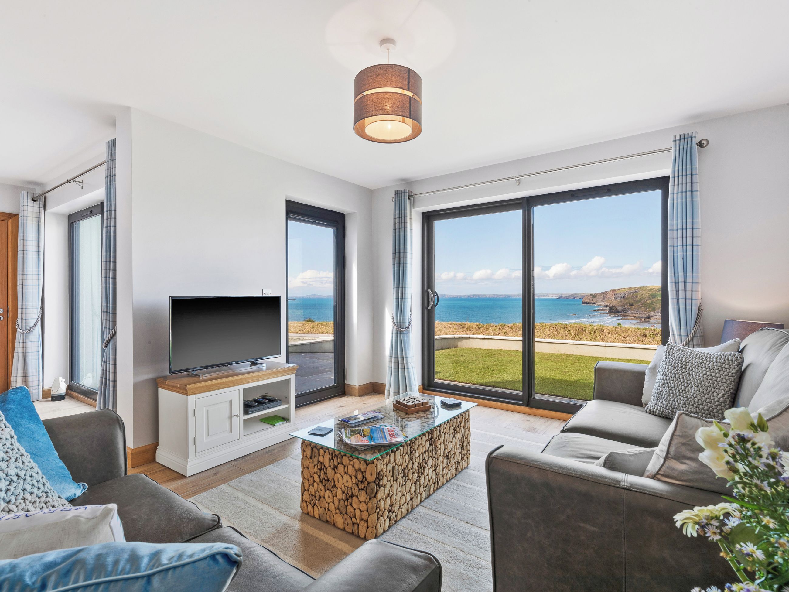 A modern living room with a television, seating area, and a large window offering a coastal view.