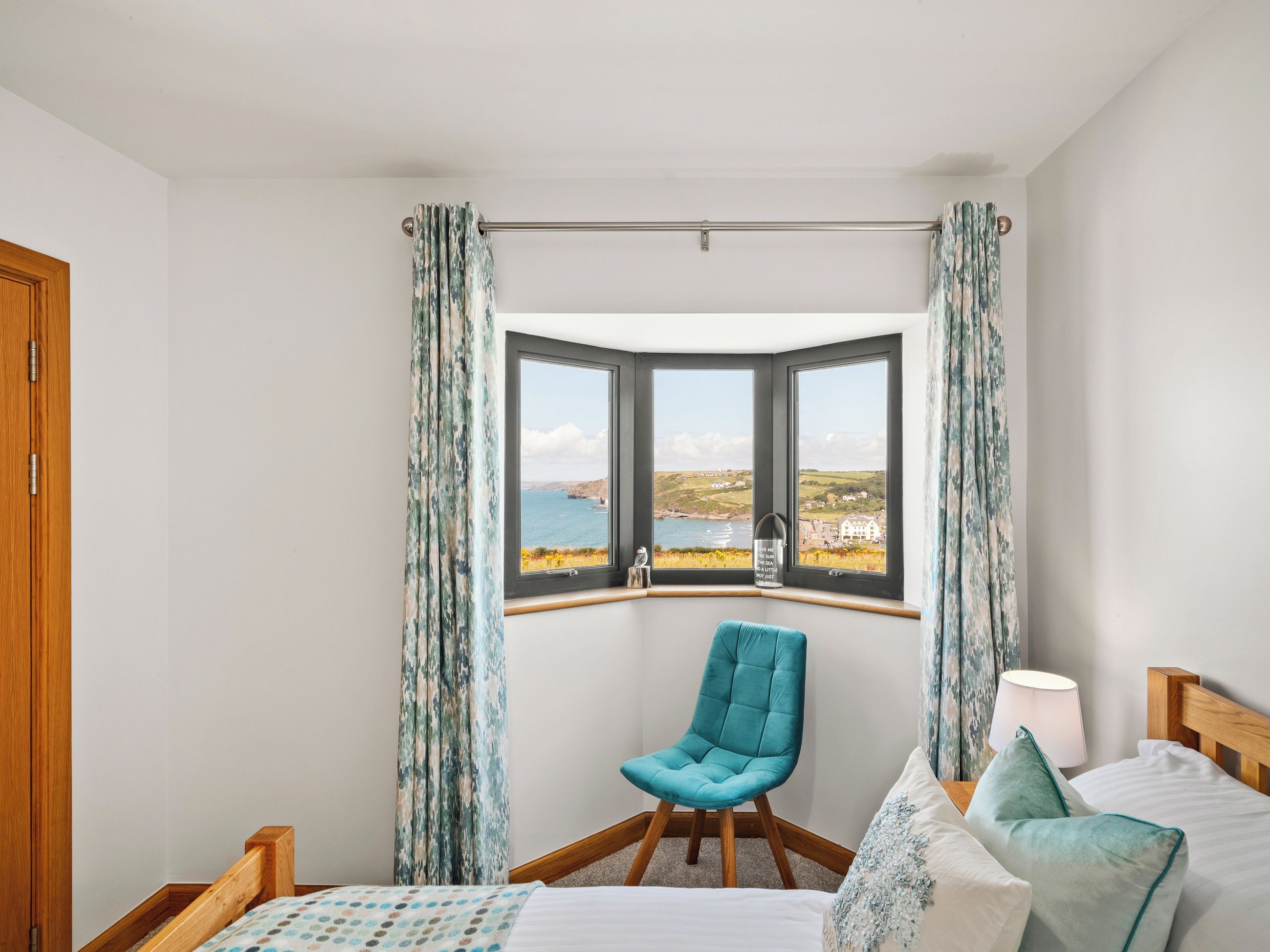 A bedroom with a bay window, a wooden bed, a teal chair, and a view of the sea.