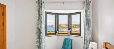 A bedroom with a bay window, a wooden bed, a teal chair, and a view of the sea.