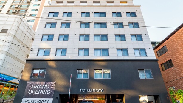 Um hotel com vários andares e fachada moderna, com uma placa anunciando a inauguração e vários carros estacionados na frente.