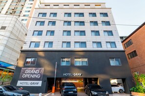 Un hotel de varios pisos con fachada moderna, un cartel que anuncia una gran inauguración y varios coches aparcados en la entrada.