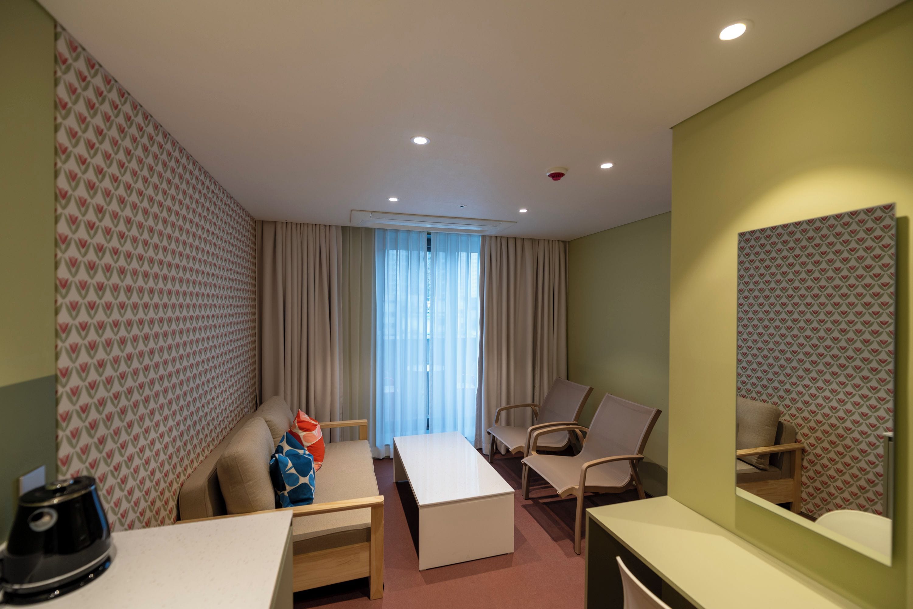 A hotel room with a bed, a desk, two chairs, a coffee maker, and a large window with curtains.