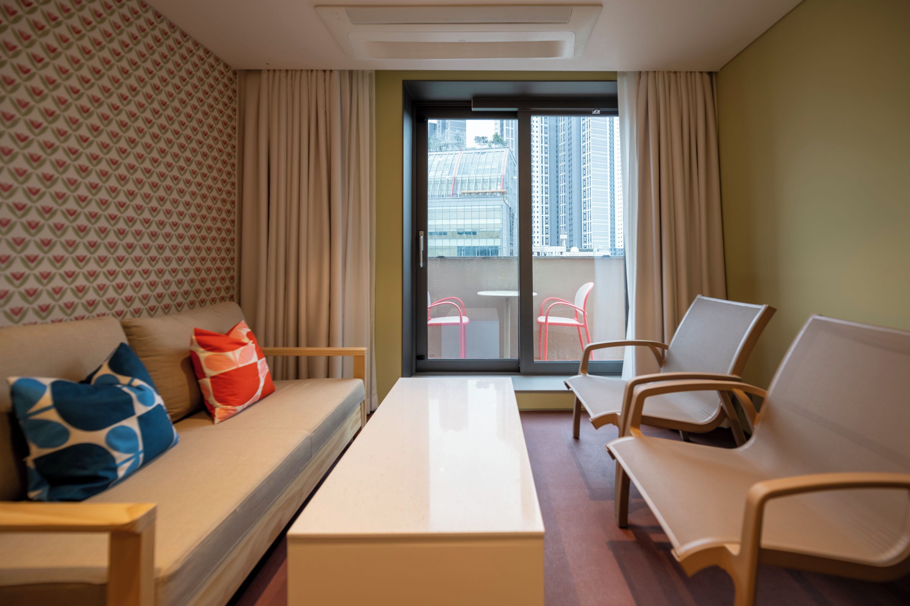 A hotel room with a sofa, two armchairs, a small table, and a balcony with a view of tall buildings.
