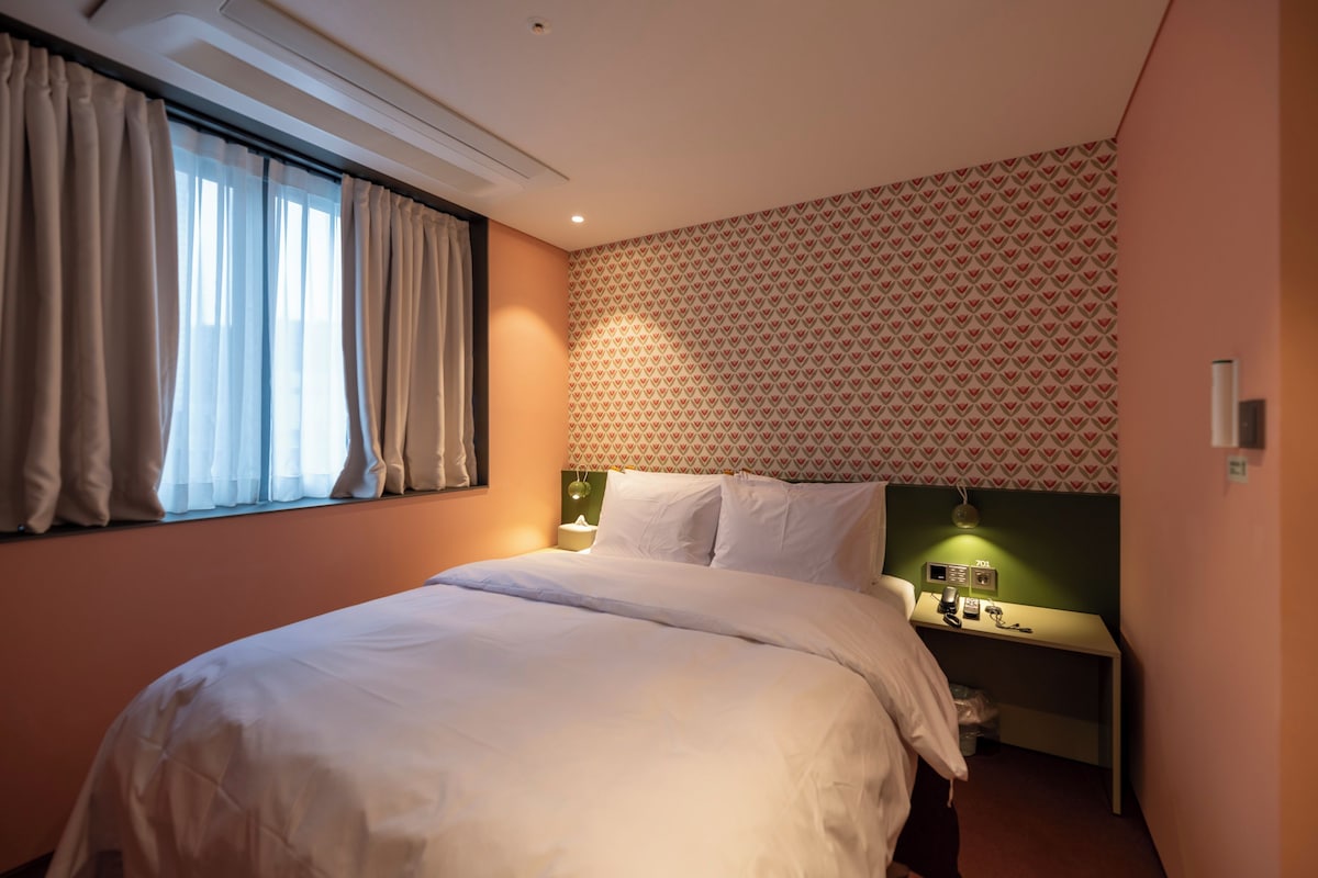 A hotel room with a bed, a bedside table with a lamp, a telephone, and a wall-mounted radio.