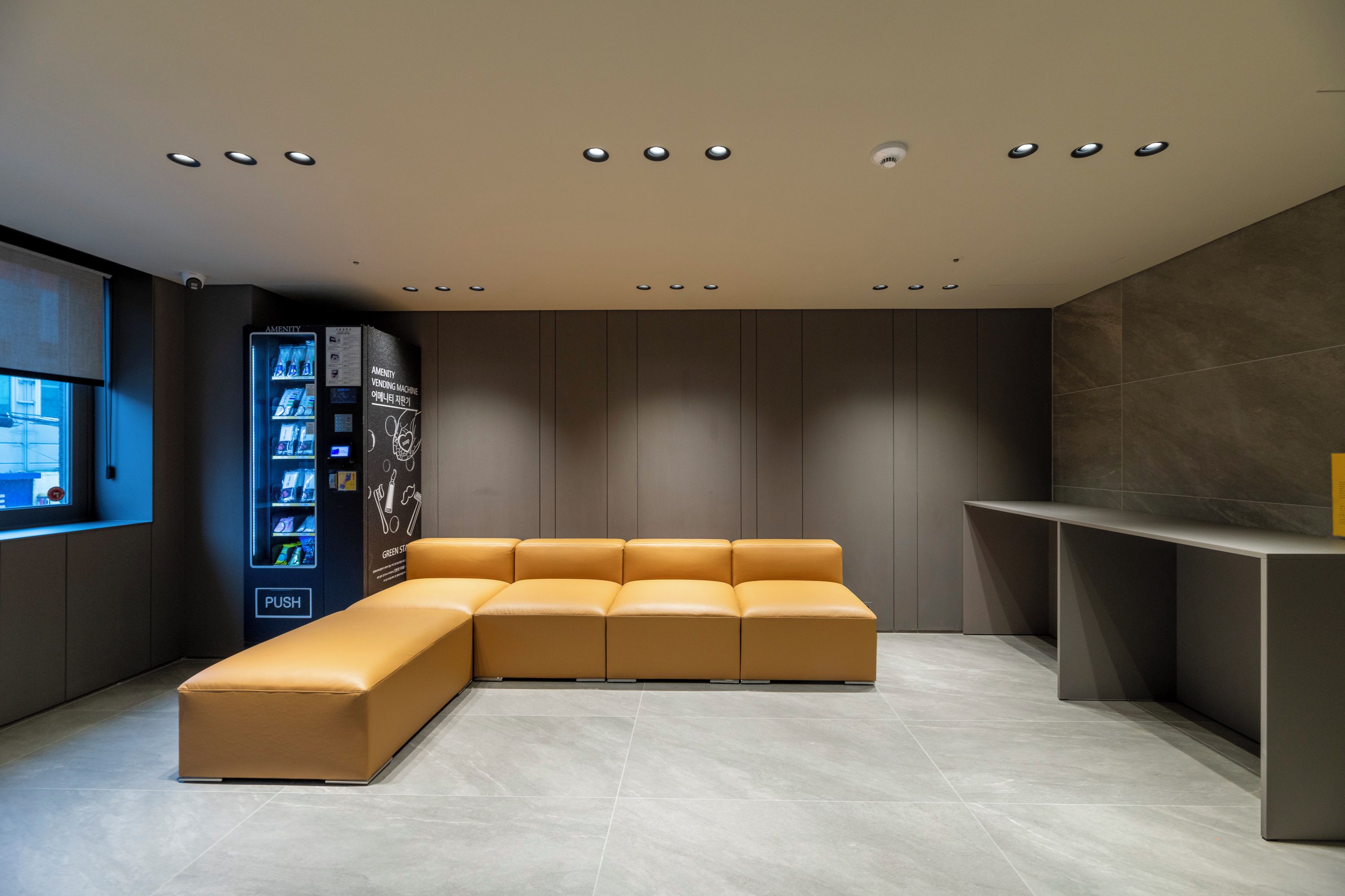 Un área de espera moderna con un sofá modular, una máquina expendedora y una pequeña recepción.