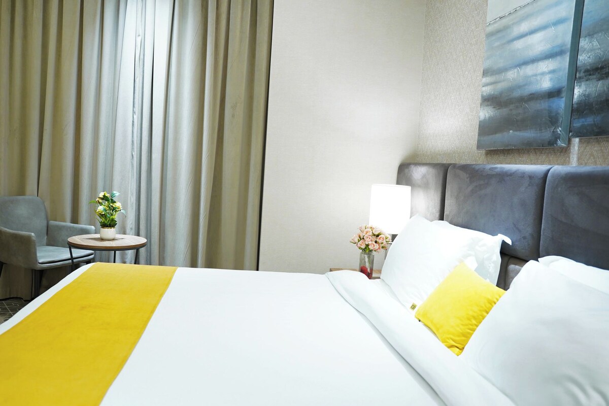 A hotel room with a bed, a chair, a small table, and a vase with flowers.