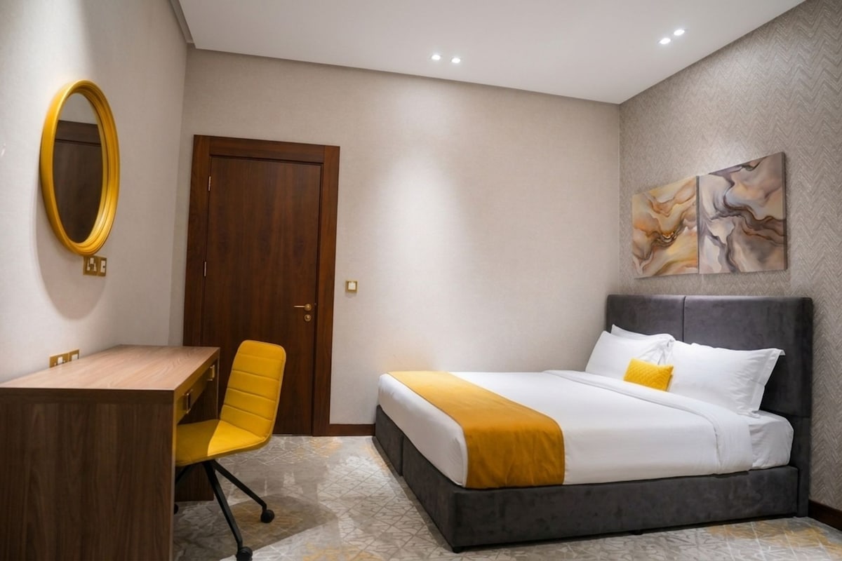 A hotel room with a bed, a desk, a chair, a mirror, and a painting on the wall.