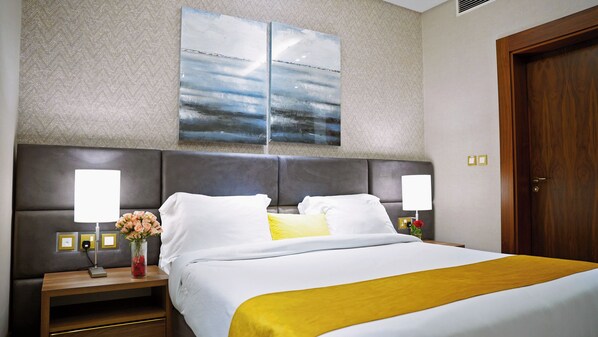 A hotel room with a large bed, two bedside tables, a nightstand, a wall-mounted artwork, a wooden door, and a wall switch. - Abraj Inn Suites Hotel (Doha)