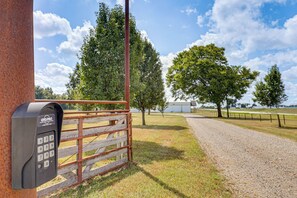 A metal gate with a keypad, a gravel path, and a fenced area with trees and a building in the background.