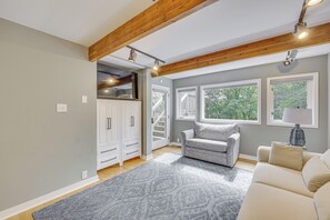 Ein modernes Wohnzimmer mit einem grauen Sofa, einem beigen Sessel und einem grauen Teppich. Weiße Schränke mit Schubladen, ein an der Wand montierter Fernseher und eine Treppe, die zu einer weiteren Ebene führen.