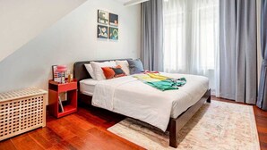 Een slaapkamer met een bed, een nachtkastje, een rode bijzettafel, een houten kist en een muur met abstracte kunst.