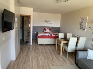 A modern living room with a sofa, a dining table with chairs, a TV mounted on the wall, and a hallway leading to a bathroom. - Directly on Lake Ossiach 1 (Steindorf am Ossiacher See)
