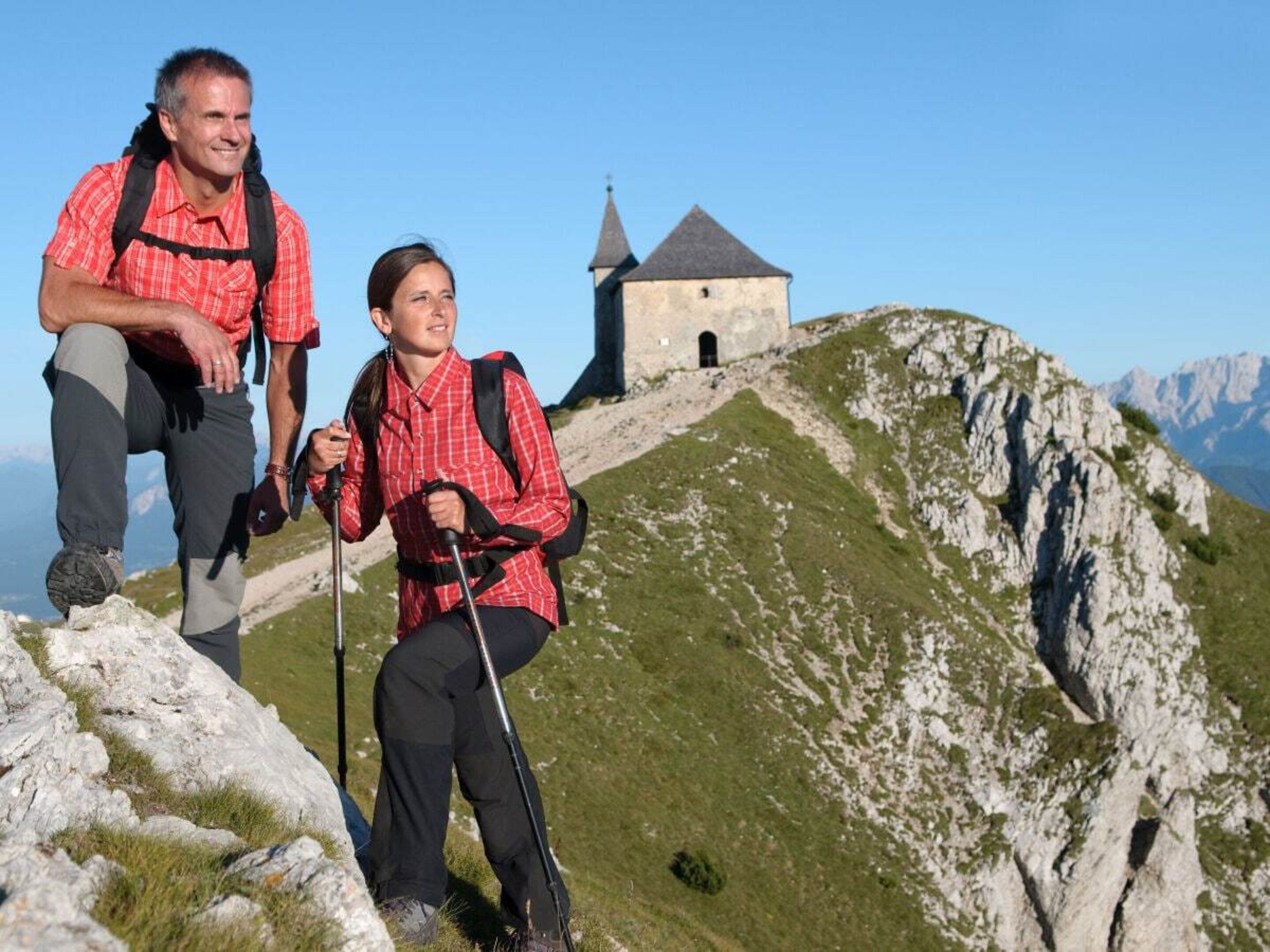Zwei Wanderer auf einem Bergpfad, im Hintergrund ein kleines Steinhaus.