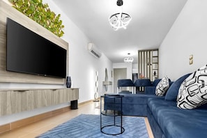 Una sala de estar moderna con un televisor de pantalla plana, un sofá azul y un comedor al fondo.
