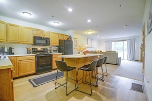 A modern kitchen with wooden cabinets, a stainless steel refrigerator, and a central island with bar stools. - Mountain-View Cedar Creek Condo w/ Trail Access (Conway)