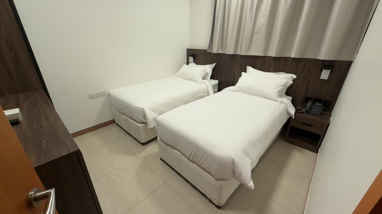 Habitación de hotel con dos camas individuales, cabecero de madera, mesita de noche con teléfono y enchufe en la pared.