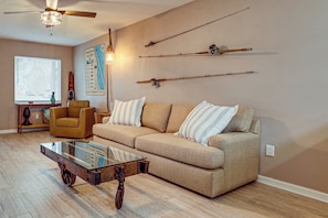 A living room with a beige sofa, a glass coffee table, a ceiling fan, and wall-mounted fishing rods. - Boat Ramp, Fishing Pier & Game Room Retreat (Edgewater)