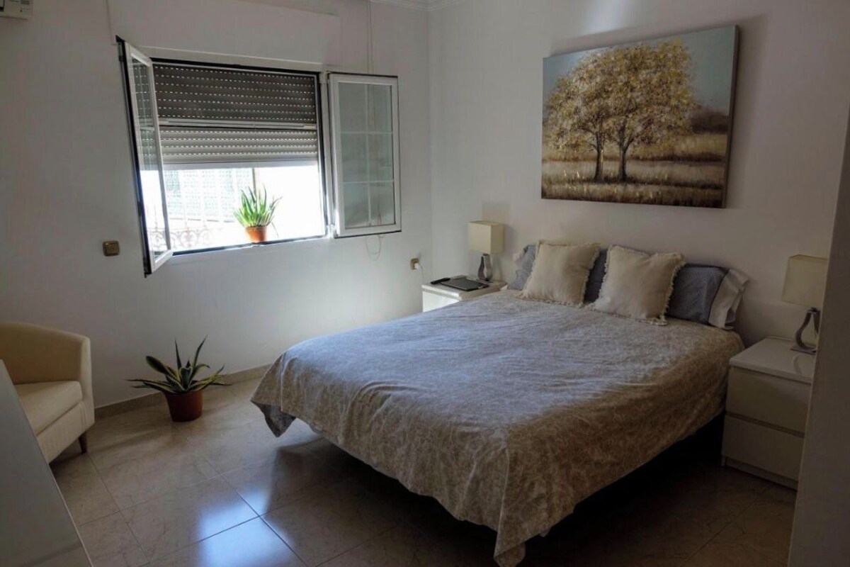 Un dormitorio con cama, ventana con persianas, mesita de noche con lámpara, una silla y una planta en maceta.