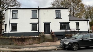 A two-story building with a black door and windows, a brick wall, and a parked car. - Standard Triple Room - Shared (Manchester)