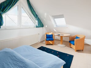 A bedroom with a bed, two wicker chairs with blue cushions, a small round table, and a window with curtains. - Holiday Apartment in Juist With Swimming Pool (Juist)