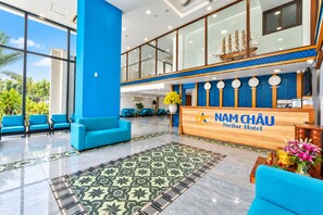A hotel lobby with a reception desk, blue seating, and a large window offering a view of greenery. - Nam Châu Stellar Hotel (Phu Quoc)