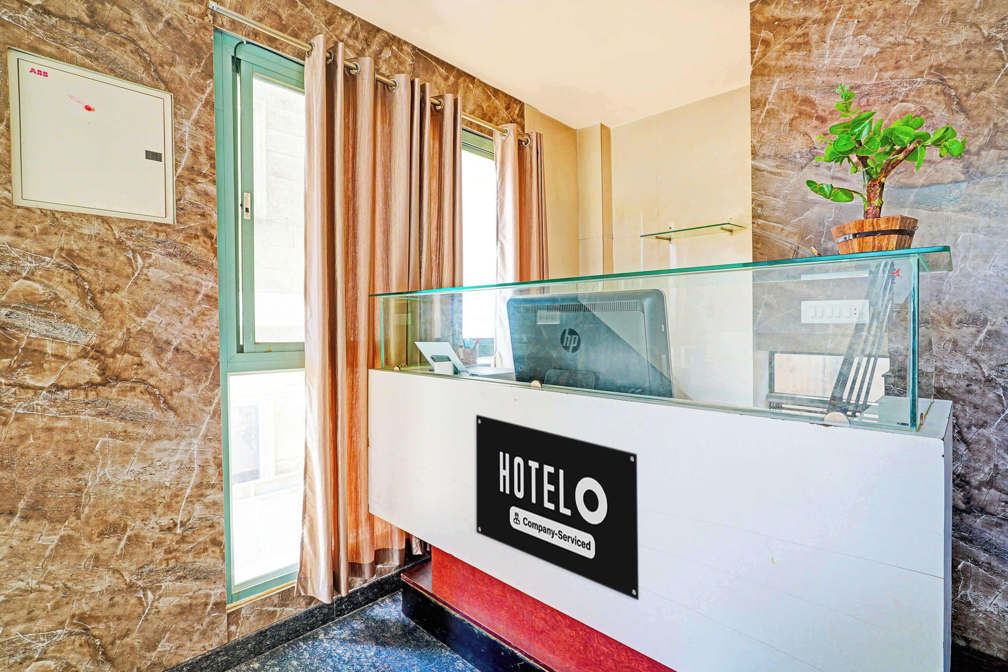 A hotel reception desk with a glass partition, a sign indicating it's company-serviced, and a potted plant on the right.
