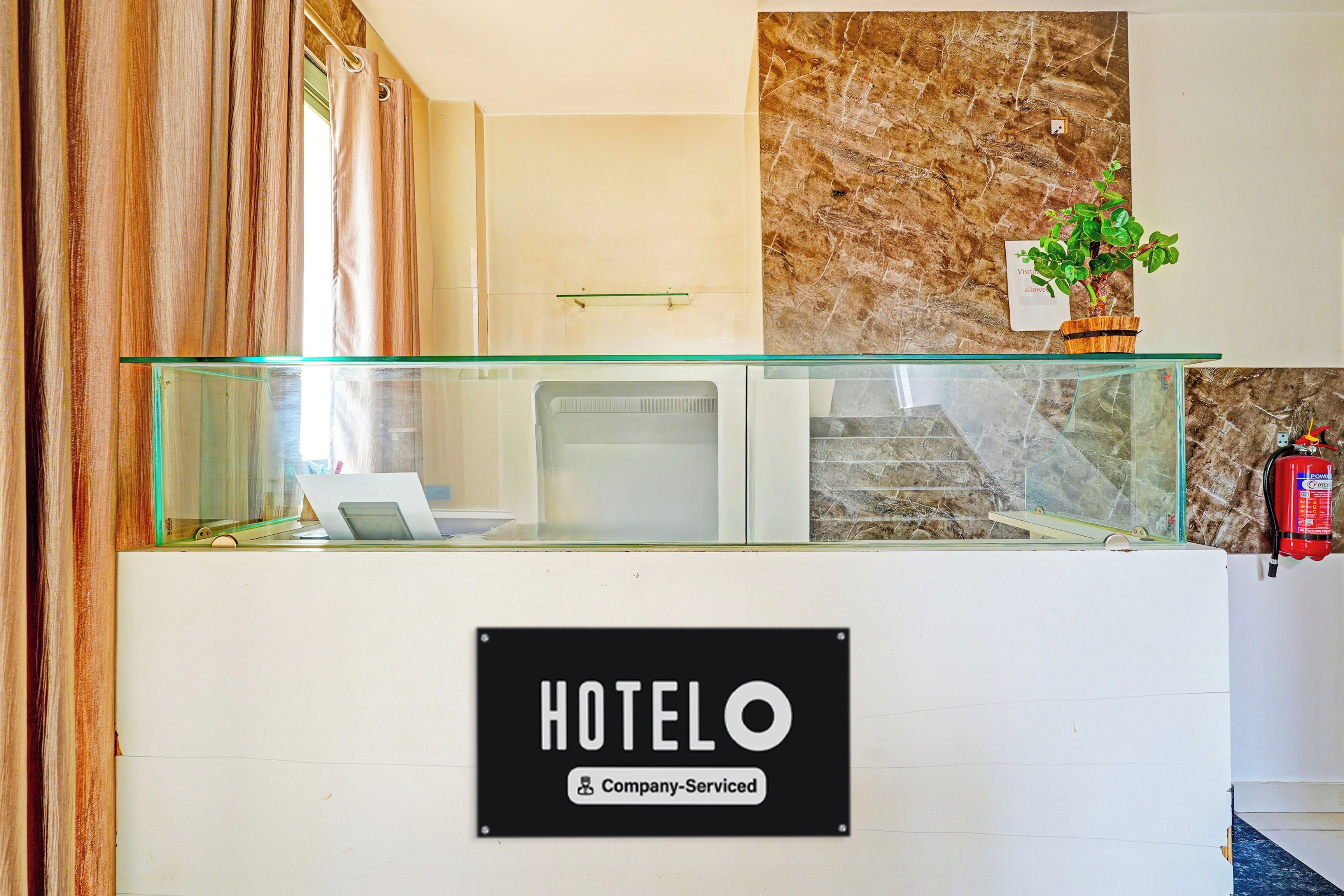 A hotel reception desk with a glass partition, a sign indicating it's a company-serviced hotel, a fire extinguisher, and a small plant on the wall.