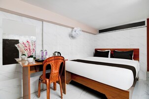 A hotel room with a bed, a desk with a chair, a mirror, and a wall-mounted fan.