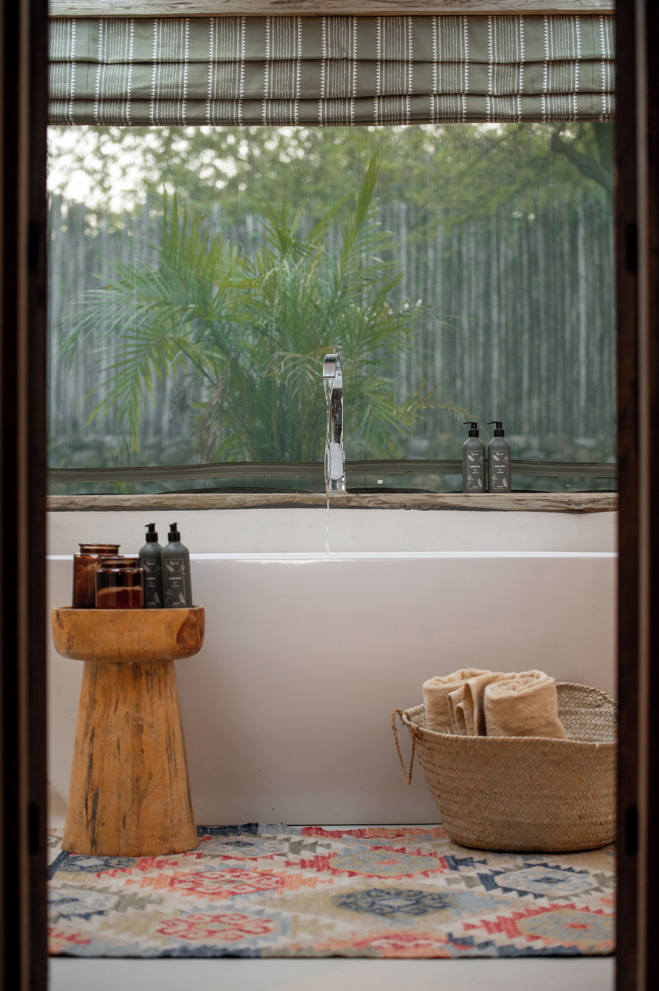 A bathroom with a bathtub, a wooden stool, a woven basket, and a window with a view of greenery.