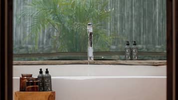 A bathroom with a bathtub, a wooden stool, a woven basket, and a window with a view of greenery.