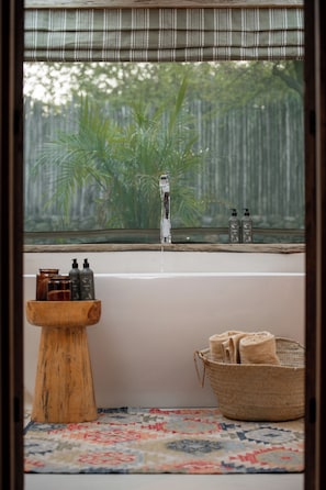 A bathroom with a bathtub, a wooden stool, a woven basket, and a window with a view of greenery.