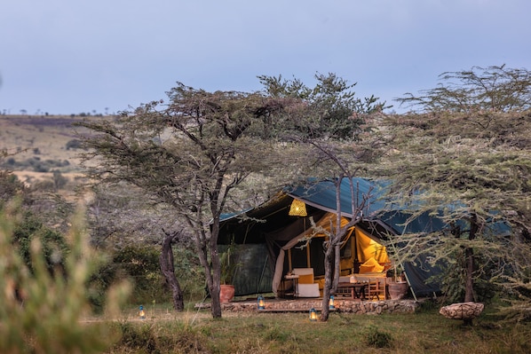A tent with a covered porch, surrounded by trees and a stone wall.