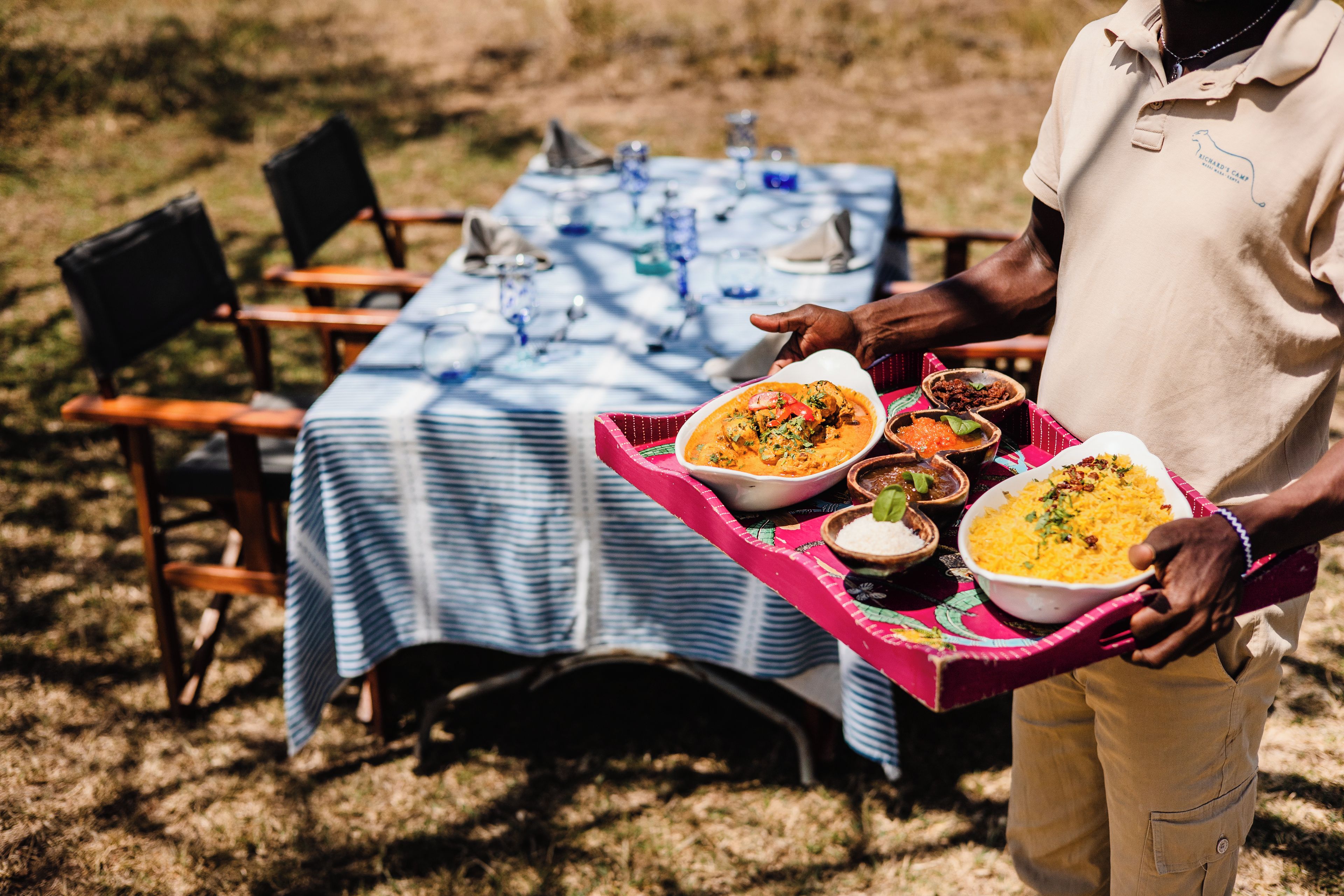 A person carrying a tray with various dishes, a table with chairs, and a blue tablecloth.