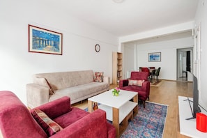 Ein modernes Wohnzimmer mit einer Couch, Sesseln, einem Couchtisch und einem Fernseher. An der Wand befinden sich ein Bücherregal und ein Gemälde.