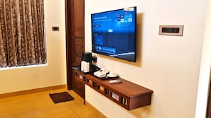 A hotel room with a flat-screen TV mounted on the wall, a wooden console table with a remote control, a tray with a coffee maker, and a window with curtains. - PL.A Resorts (Thanjavur)