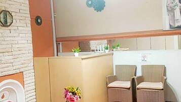 A hotel lobby with a reception desk, two wicker chairs, a red sofa, a glass coffee table, and a wall clock.