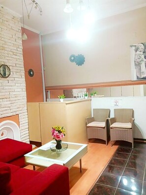 A hotel lobby with a reception desk, two wicker chairs, a red sofa, a glass coffee table, and a wall clock.