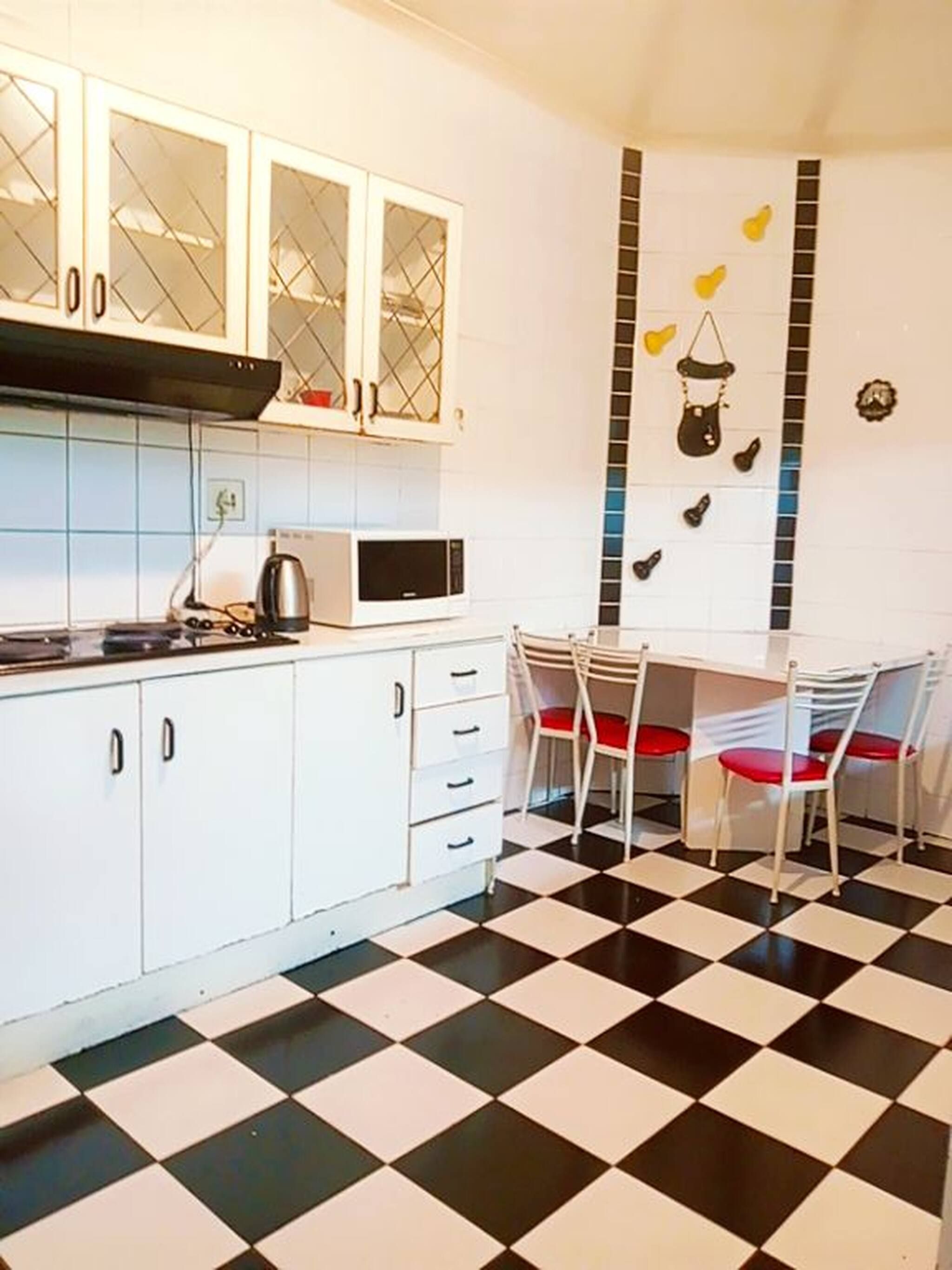 Een keuken met witte kasten, een magnetron en een gasfornuis. Er zijn twee stoelen met rode kussens en een zwart-wit geruit vloertje.