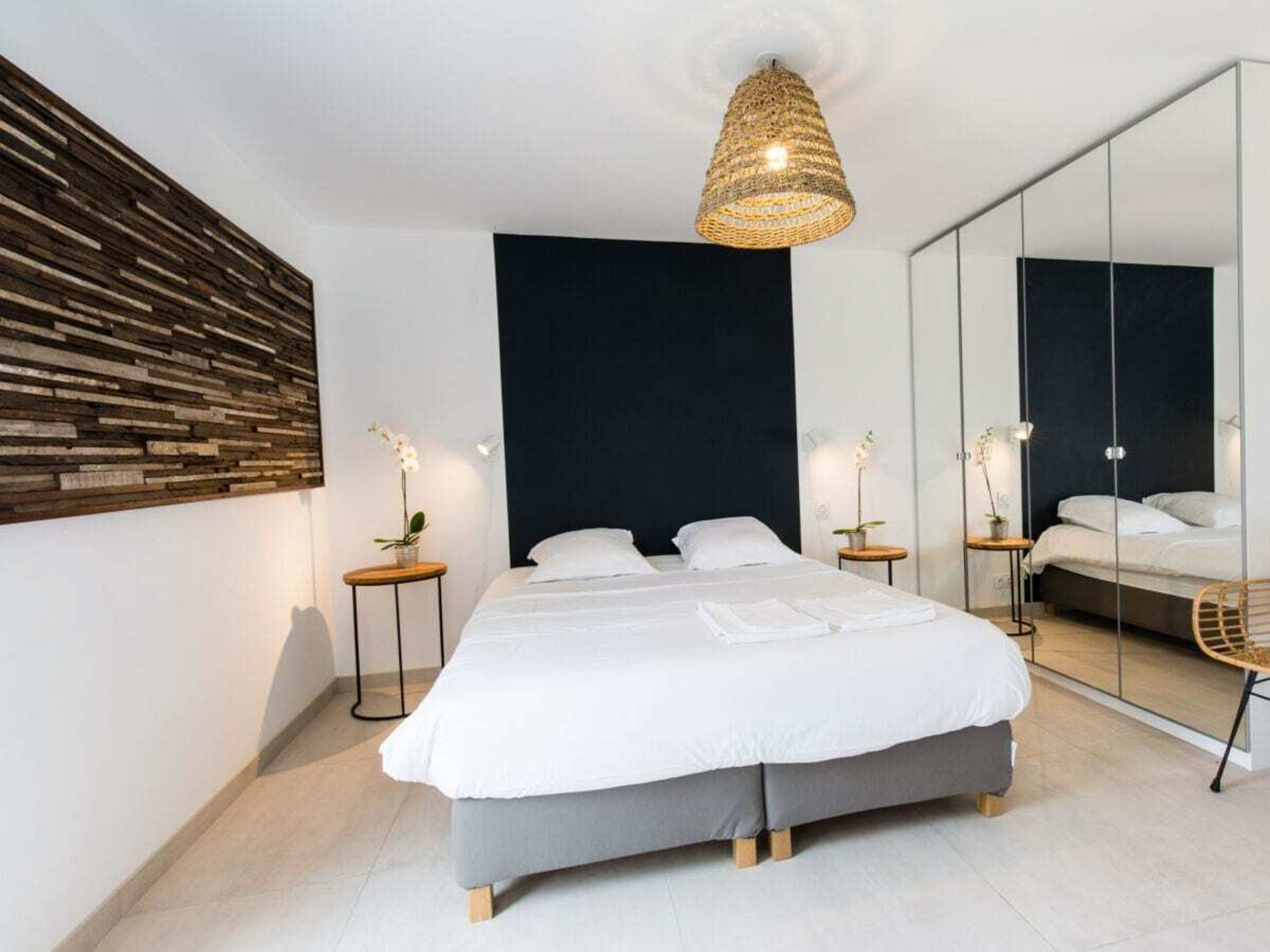 A modern bedroom with a large bed, a dark accent wall, a woven light fixture, and a full-length mirror.