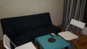 Quarto com um sofá preto, uma cadeira branca, uma mesinha com toalha de mesa verde-água e um controle remoto sobre a mesa.