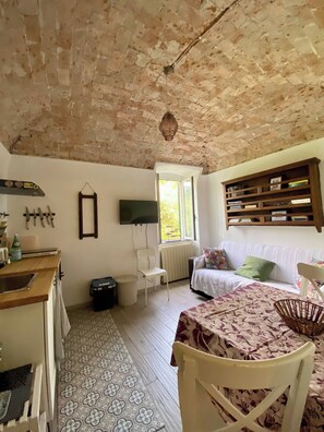 Ein Zimmer mit Ziegeldecke, einem weißen Stuhl, einem Bett mit gemustertem Überwurf, einer Holzregal mit Büchern und einem Fenster mit Blick auf Bäume.