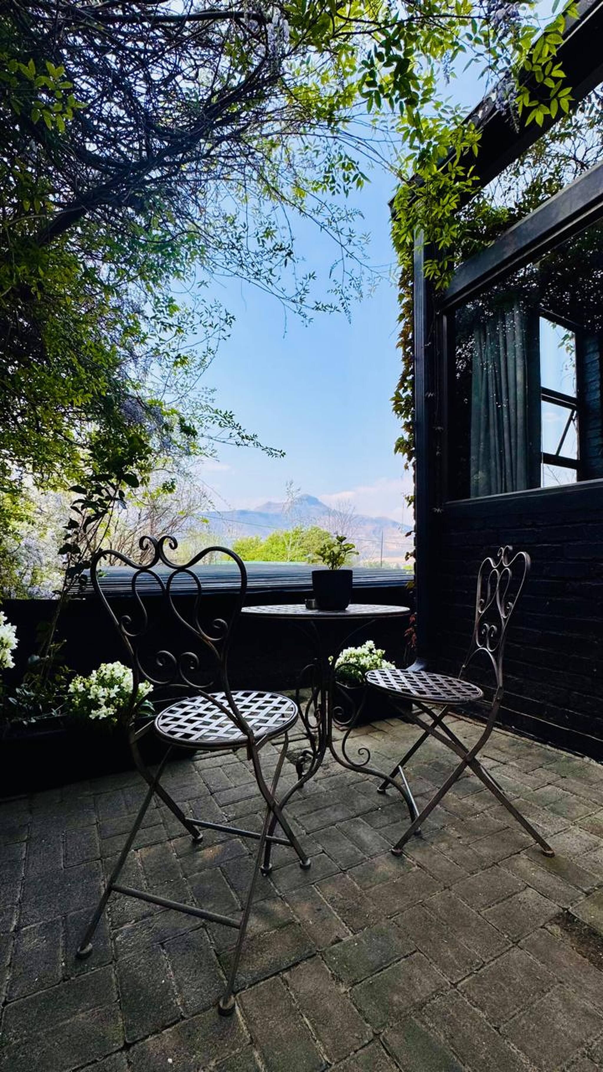 A patio with two metal chairs and a table, surrounded by greenery and a view of mountains.