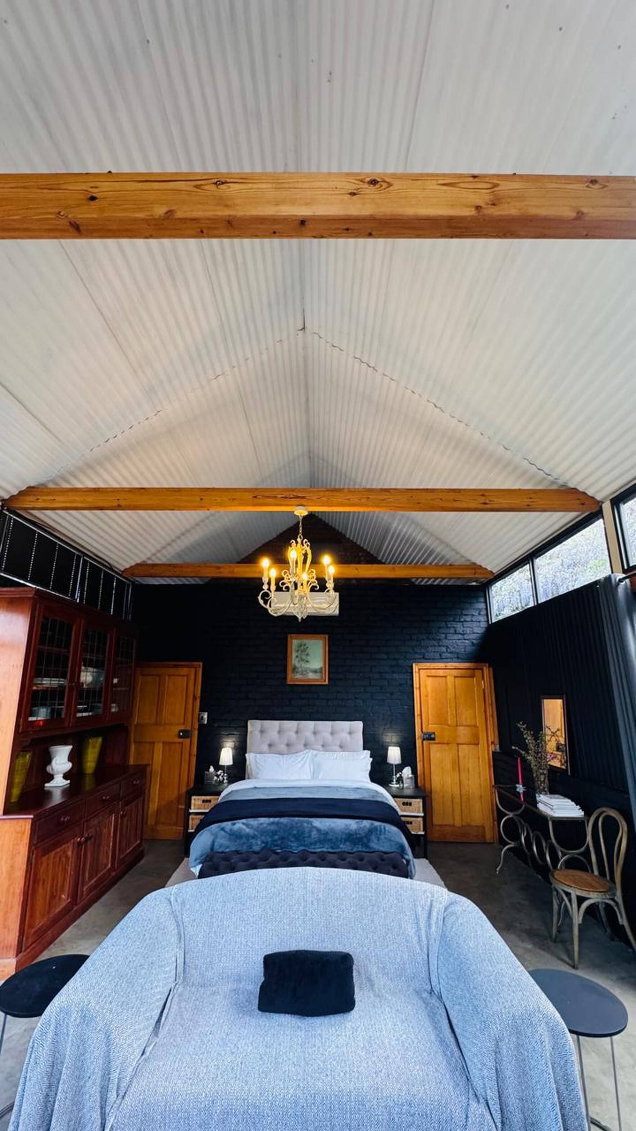 A bedroom with a large bed, a chandelier, wooden beams, a dark brick wall, and a window.